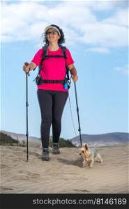 Cheerful female hiker with trekking poles and Yorkshire Terrier walking on Famara beach in summer and looking away. Traveling woman with dog on sandy beach