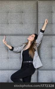 Cheerful female in smart casual clothes raising arm with clenched fist ans smiling while leaning on soft panel wall and taking selfie. Happy woman taking selfie near panel wall