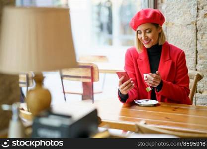 Cheerful female in stylish red beret and coat text messaging on cellphone while sitting at table with cup of tea in hands in cafeteria. Charming elegant woman surfing smartphone in cafe