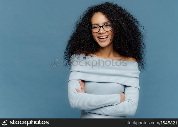 Cheerful female student has perfect mood, curly bushy hair, arms folded, looks somewhere with positive facial expression, wears blue sweater, stands indoor. People, feelings and emotions concept.