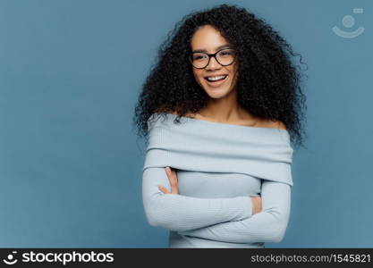 Cheerful female student has perfect mood, curly bushy hair, arms folded, looks somewhere with positive facial expression, wears blue sweater, stands indoor. People, feelings and emotions concept.