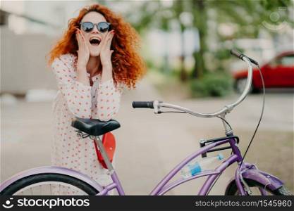 Cheerful female urban biker wears sunglasses and dress, has luxurious crisp foxy hair, keeps elbows on saddle of bicycle, spends recreation time in open air, poses over blurred street background