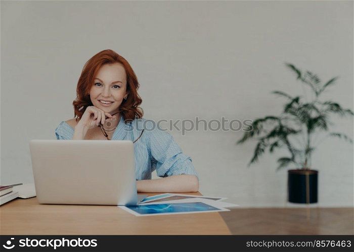 Cheerful foxy female entrepreneur chats online with business partners, works on startup project, poses at workplace, surrounded with paper documents, white wall and potted flower in background