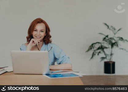 Cheerful foxy female entrepreneur chats online with business partners, works on startup project, poses at workplace, surrounded with paper documents, white wall and potted flower in background