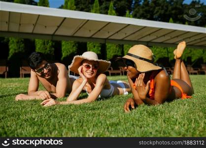 Cheerful friends rest on the grass near the pool. Happy people having fun on summer vacations, holiday party at the poolside outdoors. One man and two women are sunbathing. Cheerful friends rest on the grass near the pool