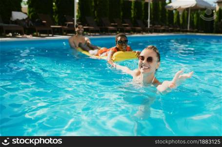 Cheerful friends swim on a mattress in the pool. Happy people having fun on summer vacations, holiday party at the poolside outdoors. One man and two women leisure at the resort. Cheerful friends swim on a mattress in the pool