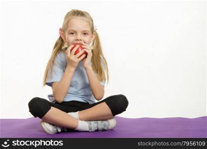 Cheerful girl eating an apple sitting on sport mat. Six year old girl Europeans engaged in physical exercises