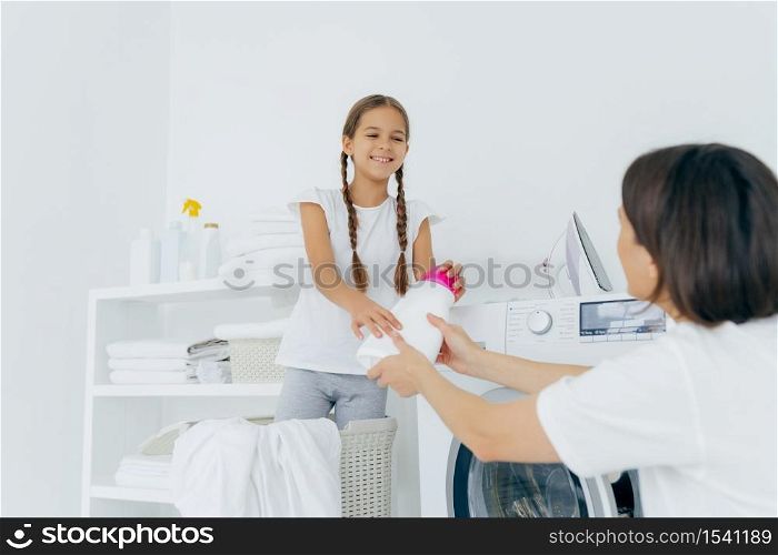Cheerful girl gives washing detergent to mother, being in laundry room, put linen into washer, wash clothes, busy with domestic everyday routine, have much work about house. Equipment for cleaning