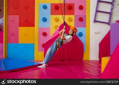 Cheerful girl in helmet poses at the climbing wall, entertainment center, young climber. Children having fun, kids spend the weekend on playground, happy childhood. Cheerful girl in helmet poses at the climbing wall