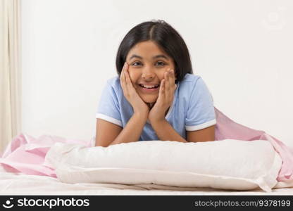 Cheerful girl looking at camera with hands on cheek while lying down on bed 