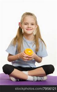 Cheerful girl sitting with orange on the sports rug. Six year old girl Europeans engaged in physical exercises