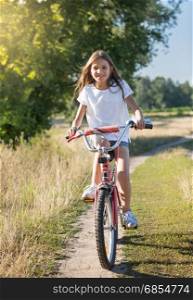 Cheerful girl with long hair riding her bicycle on dirt road at meadow