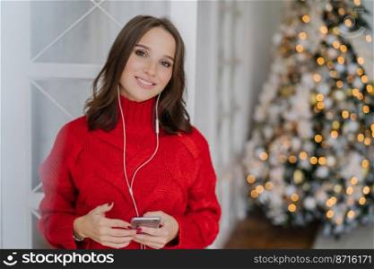 Cheerful good looking woman has toothy smile, enjoys nice track in earphones, updates playlist on smart phone, dressed in warm knitted sweater, poses against decorated Christmas tree in background