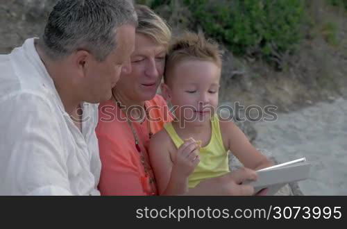 Cheerful grandfather, grandmother and little grandchild with tablet PC outdoor. They watching cartoons and boy eating cookie