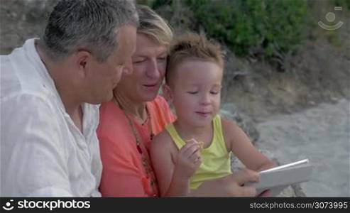 Cheerful grandfather, grandmother and little grandchild with tablet PC outdoor. They watching cartoons and boy eating cookie