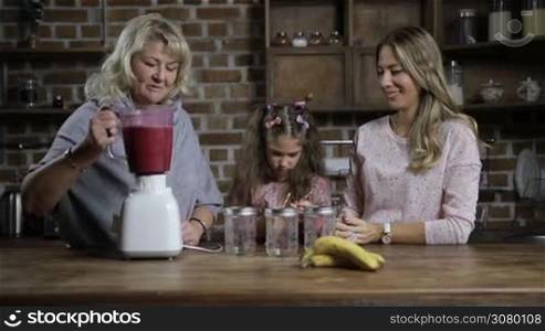 Cheerful grandmother pouring fresh blended berry smoothie into mason jars while family sharing healthy breakfast together in the kitchen. Multi generation family enjoying tasty healthy smoothie at home. Dooly shot. Slow motion.