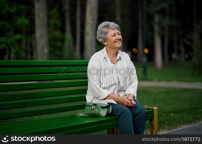 Cheerful granny leisures on the bench in summer park. Aged people lifestyle. Pretty grandmother having fun outdoors, old female person on nature. Cheerful granny leisures on bench in summer park