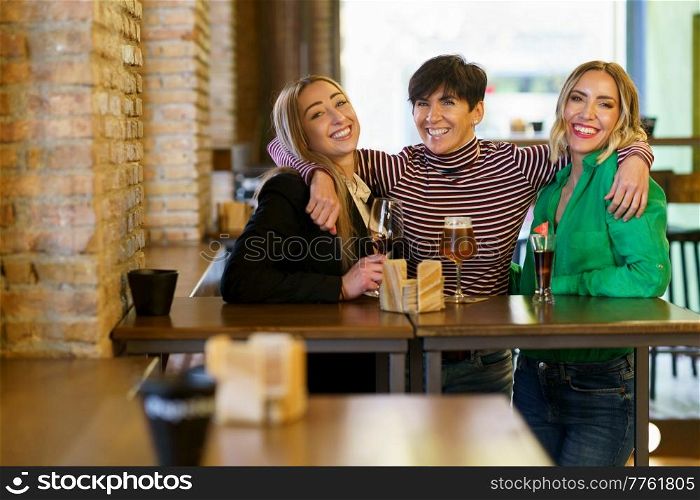 Cheerful group of female friends looking at camera with smile and embracing girlfriends behind table with alcohol during meeting in pub. Cheerful girlfriends hugging during party in bar