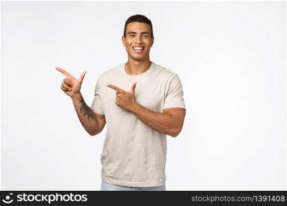 Cheerful handsome muscline man with tattoo on arm, tan, wearing white t-shirt, pointing left and smiling camera happy, introduce product or promo, recommend new place hang out, studio background.. Cheerful handsome muscline man with tattoo on arm, tan, wearing white t-shirt, pointing left and smiling camera happy, introduce product or promo, recommend new place hang out, studio background