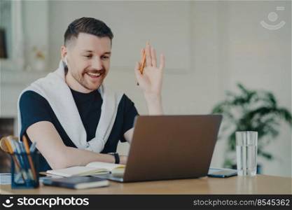 Cheerful handsome young businessman or male freelancer wearing earphones waving at webcam on laptop and saying hello to his colleagues or client during video conference, working at home office. Happy businessman greeting colleague during video conference