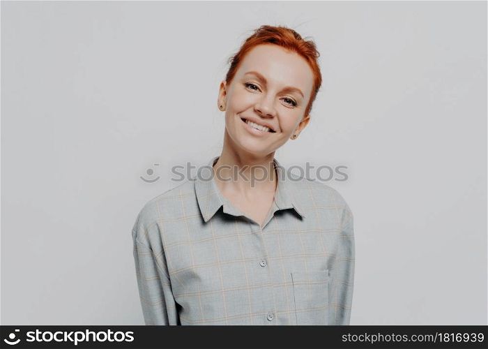 Cheerful happy beautiful red haired woman with beaming smile posing isolated on grey studio background, dressed in casual checkered shirt, expressing positivity and happiness. Face expressions concept. Cheerful beautiful red haired woman with beaming smile posing isolated on grey studio background