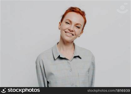 Cheerful happy beautiful red haired woman with beaming smile posing isolated on grey studio background, dressed in casual checkered shirt, expressing positivity and happiness. Face expressions concept. Cheerful beautiful red haired woman with beaming smile posing isolated on grey studio background