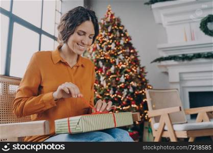 Cheerful happy European woman unpacking Xmas gifts at home, anticipating joyful mood of upcoming winter holidays, smiling Italian female sitting in room with decorated New Year tree. Happy European woman unpacking Xmas gifts while sitting in room with decorated New Year tree