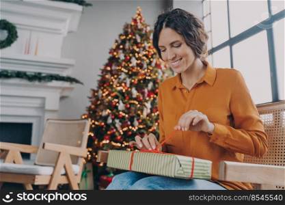 Cheerful happy European woman unpacking Xmas gifts at home, anticipating joyful mood of upcoming winter holidays, smiling Italian female sitting in room with decorated New Year tree. Happy European woman unpacking Xmas gifts while sitting in room with decorated New Year tree