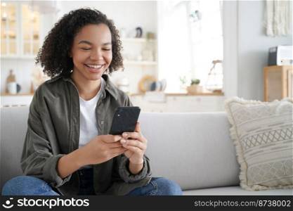 Cheerful happy mixed race teen girl reading message with good news on smartphone, smiling laughing young lady watching funny videos in social networks, sitting on sofa, using mobile apps on phone.. Happy mixed race girl uses mobile phone apps watching funny video in social networks sitting on sofa