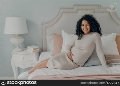 Cheerful happy pregnant mixed race future mom with curly hair relaxing on big comfy bed after long trip to shopping mall, being on last months of pregnancy, holding her belly with one hand and smiling. Smiling young pregnant mixed race lady resting on bed and enjoying last months of pregnancy