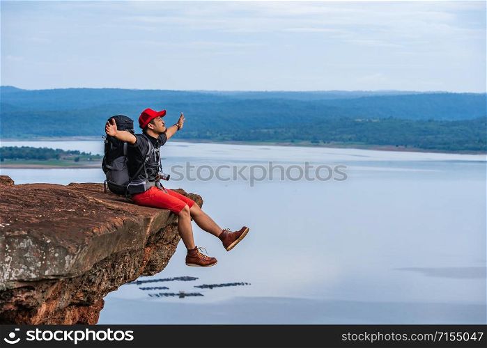 cheerful hiker man sitting and gesture raised arms on the edge of cliff, on a top of the rock mountain
