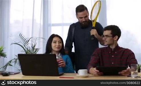 Cheerful hipster businessman with beard holding tennis racket in creative office and going to play tennis. Sporty male tennis player going for training as his coworkers working in modern office.