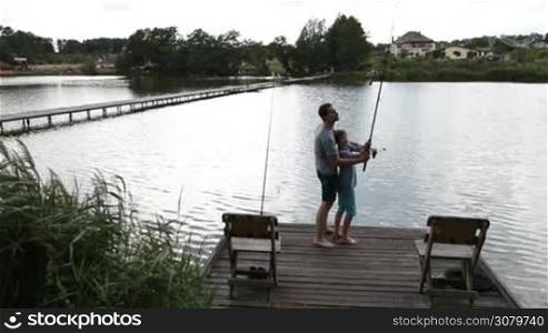 Cheerful hipster father and teenage son standing barefoot on wooden pier andn agling with fishing rods at tranquil freshwater pond. Back view. Young dad helping his boy to throw spinning rod and reel while fishing together on the lake in summer.
