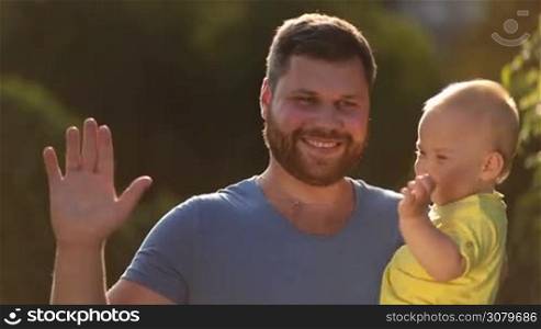 Cheerful hipster father with beard carrying cute toddler baby boy, waving hello with hands and smiling in summer park in glow of beautiful sunset. Portrait of happy dad and infant son posing in nature, looking at camera with toothy smile.