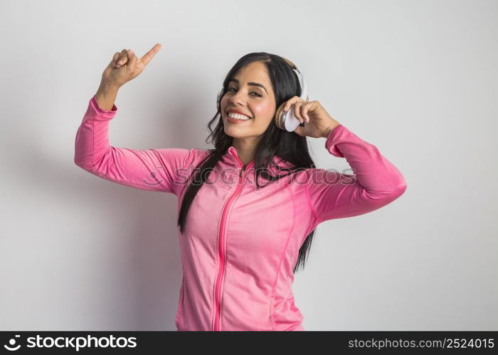 Cheerful Hispanic female model in sportswear pointing up while listening to songs in wireless headphones in studio against white background. Smiling ethnic woman in sports jacket listening to music