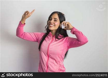 Cheerful Hispanic female model in sportswear pointing up while listening to songs in wireless headphones in studio against white background. Smiling ethnic woman in sports jacket listening to music