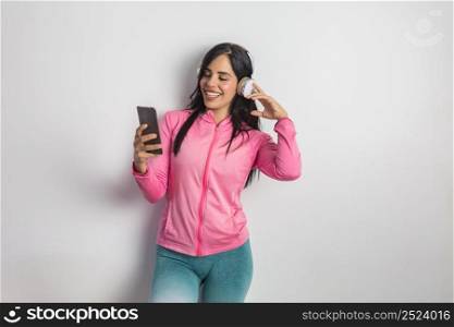 Cheerful Hispanic sportswoman in activewear searching playlist for training on cellphone while enjoying music in headphones in studio against white background. Smiling ethnic woman using smartphone and listening to music