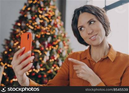 Cheerful hispanic woman making selfie on her mobile while sitting in room against backdrop of beautiful bright colored festive Xmas tree at home. Female making video call during winter holidays. Happy italian female making video call during winter holidays, talking with family online