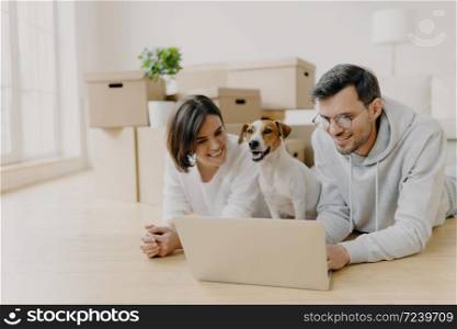 Cheerful husband and wife watch film online on laptop, rest on floor, relax and talk, their domestic animal poses between, relocate in new home, pose in spacious living room with unpacked boxes