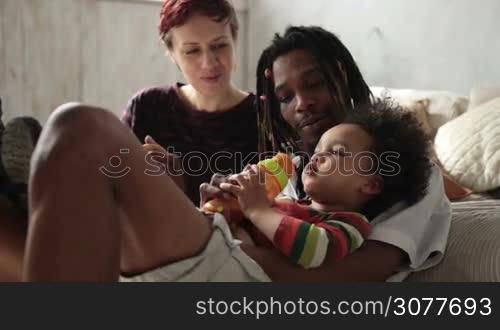 Cheerful interracial family with mixed race little son spending leisure at home. Stylish african american father with dreadlocks holding his cute child while toddler boy drinking from feeding bottle. Slow motion.