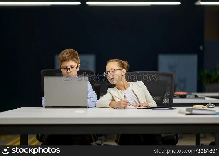 Cheerful little boy in suit talking with young girl in formalwear while sitting at working place. Boy in suit talking girl in formalwear