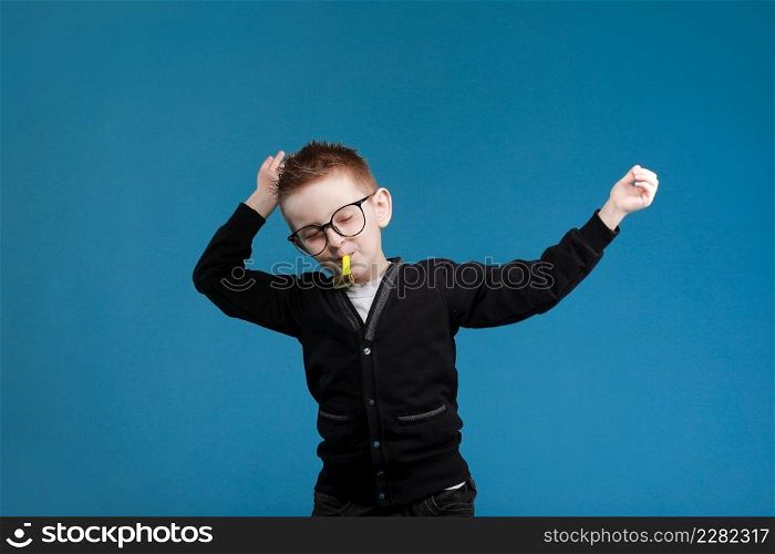 Cheerful little child boy 7s years old in glasses blowing festive pipe at birthday party isolated on blue background. children studio portrait. Childhood lifestyle concept.. Cheerful little child boy 7s years old in glasses blowing festive pipe at birthday party isolated on blue background. children studio portrait. Childhood lifestyle concept