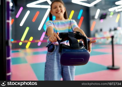 Cheerful little girl holds virtual reality glasses. Kid playing 3d video game in entertainment center. Children having fun in vr, child spend the weekend on playground, happy childhood. Cheerful little girl holds virtual reality glasses