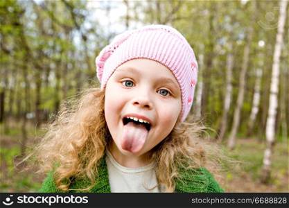 Cheerful little girl in a pink cap puts out the tongue in forest