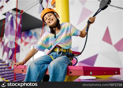 Cheerful little girl in helmet climbs on the ropes on zip line in entertainment center. Children having fun in climbing area, kids spend the weekend on playground, happy childhood. Cheerful little girl in helmet climbs on the ropes