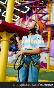 Cheerful little girl in helmet poses on zip line in entertainment center. Children having fun in climbing area, kids spend the weekend on playground, happy childhood. Cheerful little girl in helmet poses on zip line