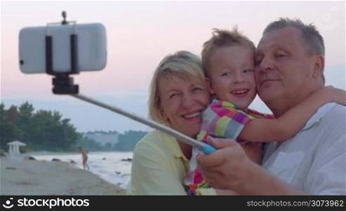 Cheerful little grandson and grandparents making selfie picture with mobile phone and monopod on the coast. They exchanging kisses to express love