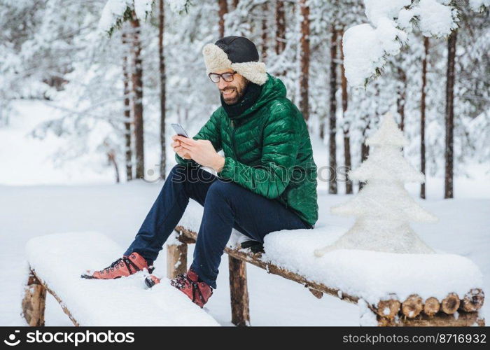 Cheerful male enjoys communication on smart phone and loneliness, calm atmosphere, sits on wooden bench covered with snow in winter forest, happy to recieve message from close friend