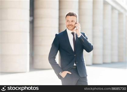 Cheerful male entrepreneur has phone conversation, looks confidently and happily aside, dressed in formal clothing, has talk with operator or business partner, keeps hand in pocket of formal suit