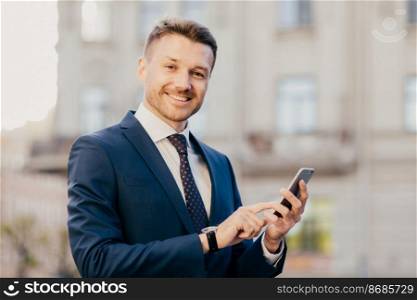 Cheerful male financier holds modern cell phone chats with partners, arrange business meeting, poses outdoors, has happy expression, dressed in black elegant suit. Successful businessman reads message